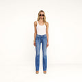 A woman stands facing forward against a plain white background with her arms relaxed by her sides, wearing sunglasses, a white tank top, beige shoes, and the Selma Bootcut Jean in medium wash stretch denim.