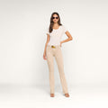 A woman wearing sunglasses, a white short-sleeve top, and heeled sandals stands against a plain light background with one hand on her hip. She pairs her outfit with the Selma Bootcut Jean, featuring a contoured waistband and belt.