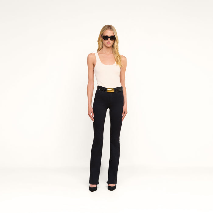 A woman with long blonde hair wears sunglasses, a white tank top, and the Selma Bootcut Jean in true black with a belt and black heels, standing against a plain white background.