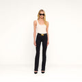 A woman with long blonde hair wears sunglasses, a white tank top, and the Selma Bootcut Jean in true black with a belt and black heels, standing against a plain white background.