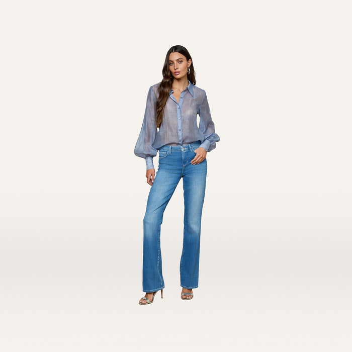 A woman with long, wavy hair stands in a relaxed pose against a plain white background, wearing the Selma Bootcut Jean with a contoured waistband, styled with a sheer blue blouse and heeled sandals, and one hand in her pocket.