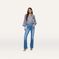 A woman with long, wavy hair stands in a relaxed pose against a plain white background, wearing the Selma Bootcut Jean with a contoured waistband, styled with a sheer blue blouse and heeled sandals, and one hand in her pocket.