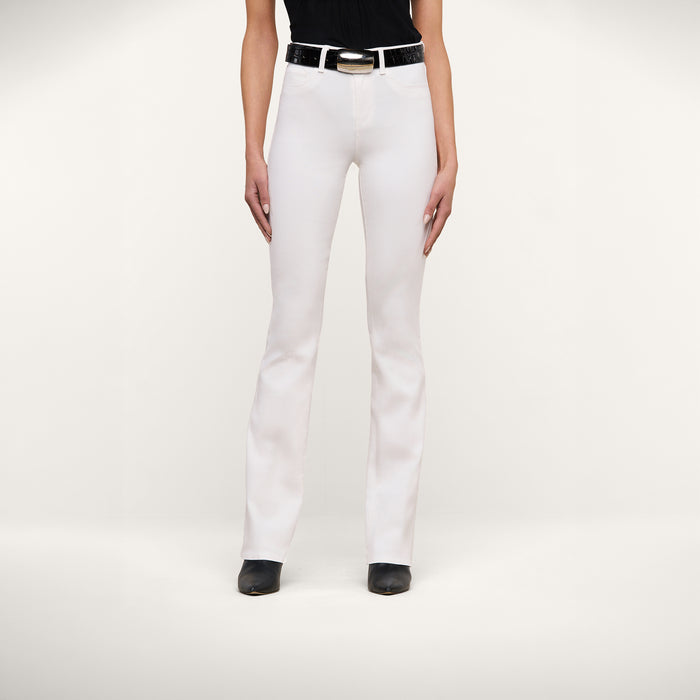 A person stands facing forward, wearing Selma Bootcut Jean in white stretch denim with a contoured waistband, paired with a black belt, black top, and black shoes. The background is plain white, and the person’s face is not visible.