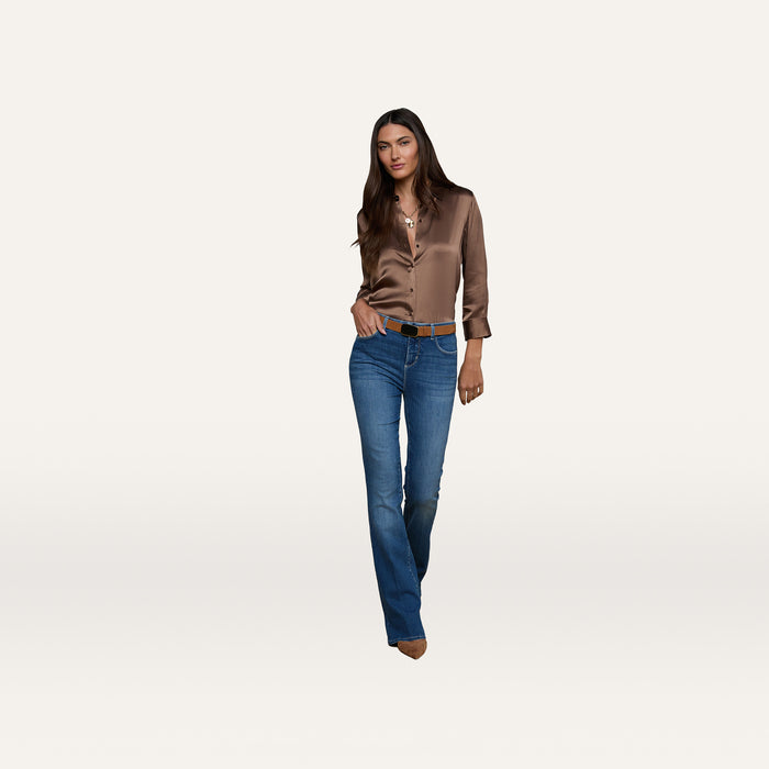 A woman stands against a plain white background, wearing dark wash Selma Bootcut Jeans made from super stretch denim, paired with a shiny brown blouse and brown pointed shoes. She has long dark hair and a neutral expression, with one hand in her pocket.