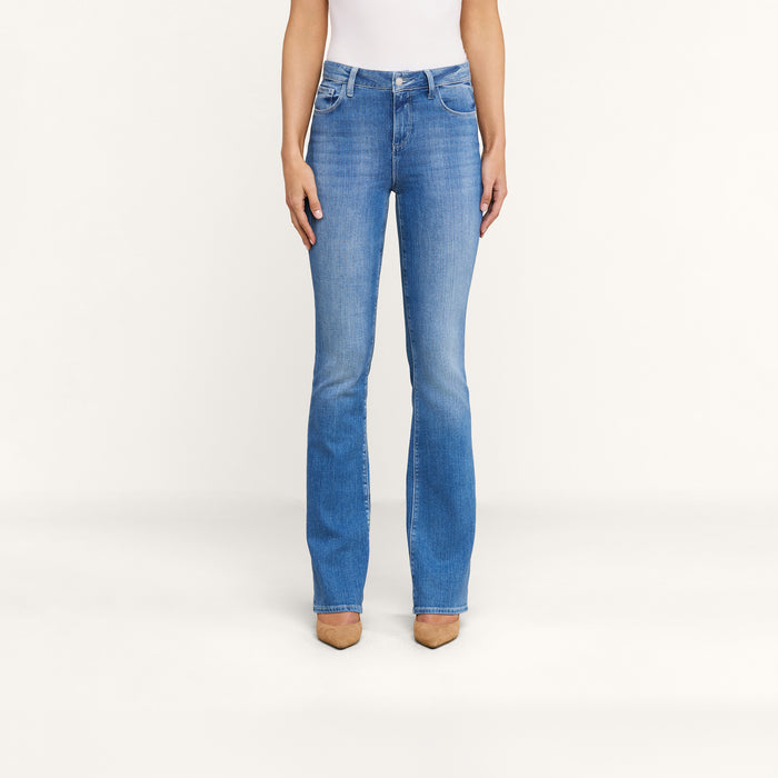 A person models the Selma Bootcut Jean in a medium wash stretch denim, paired with a white top and tan pointed-toe shoes. The image shows only the lower half of their body against a plain white background.