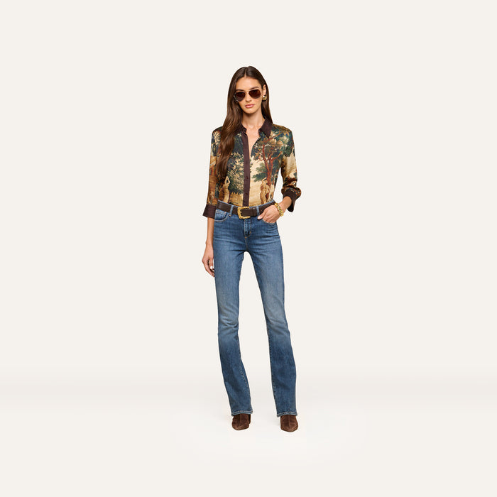 A woman stands confidently against a plain light background, wearing sunglasses, a patterned long-sleeve shirt, a belt, brown boots, and the Selma Bootcut Jean.
