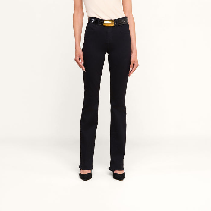 A person wearing the Selma Bootcut Jean in true black, paired with a light top, black heels, and a black belt featuring a large gold buckle stands against a plain white background. Only the lower half of the body is visible.