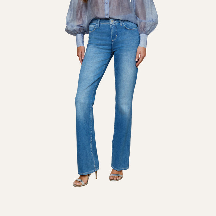 A person wearing the Selma Bootcut Jean in light blue stretch denim with a contoured waistband, paired with a sheer blue blouse with puffy sleeves and beige heeled sandals, standing against a plain background.