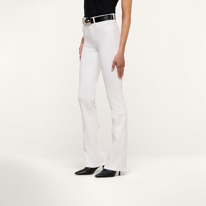 A person wearing the Selma Bootcut Jean—white stretch denim pants with a contoured waistband—paired with a black belt, black top, and black heeled shoes stands against a plain white background, their left hand resting at their side.