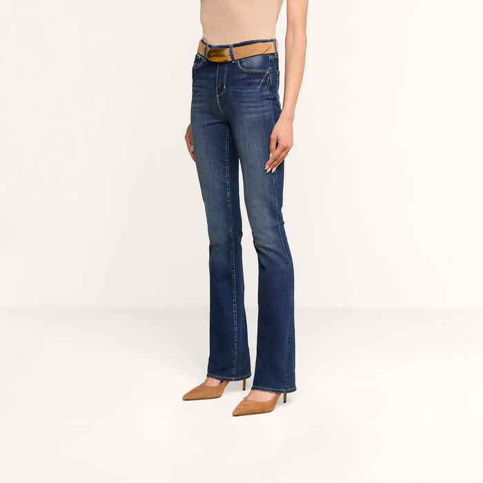 Wearing the Selma Bootcut Jean in fitted blue stretch denim, a brown belt, a beige sleeveless top, and tan pointed-toe heels, the person stands against a plain white background.