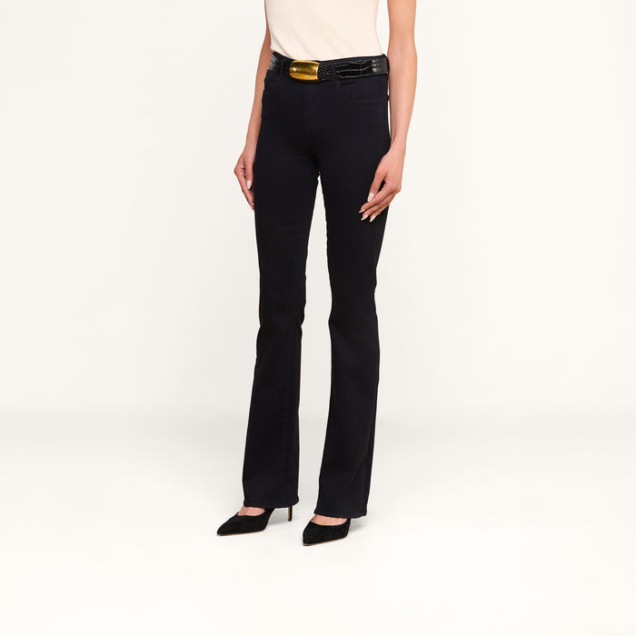 Wearing the Selma Bootcut Jean in true black with a high-rise, super stretch fit, a sleeveless white top, a black belt featuring a large gold buckle, and black pointed heels, a person stands against a plain white background.