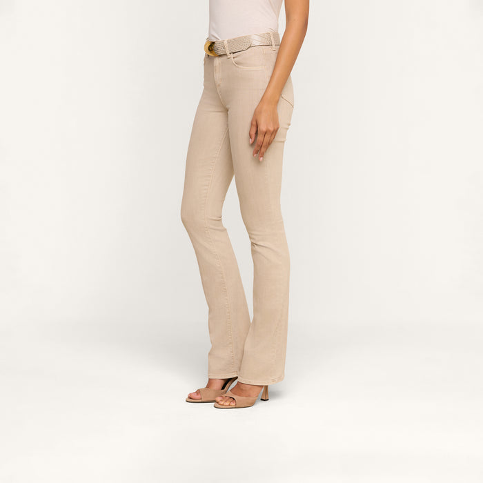 A woman stands in profile against a plain white background, wearing the Selma Bootcut Jean in beige with a contoured waistband, paired with a white top, matching belt, and open-toed heels.