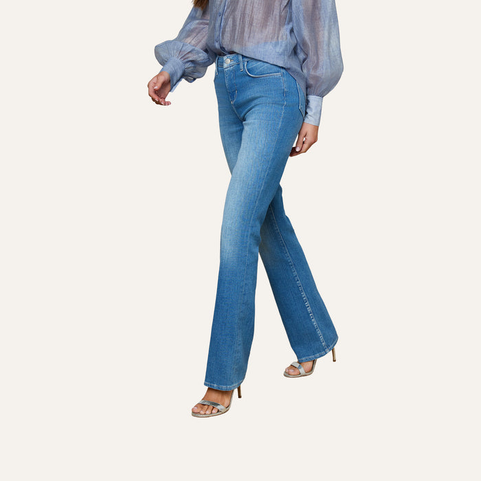 A woman wearing the Selma Bootcut Jean in light blue stretch denim, paired with a sheer blue long-sleeve blouse and silver high-heeled sandals, walks against a plain background.