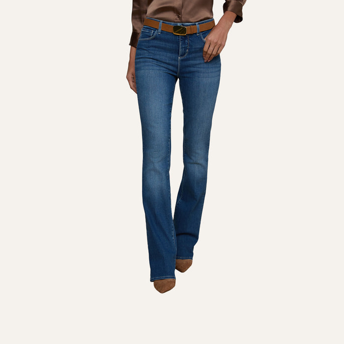 A person wearing the Selma Bootcut Jean in blue, crafted from super stretch denim, paired with a brown belt, brown pointed shoes, and a brown satin long-sleeve top. The background is plain and light-colored.