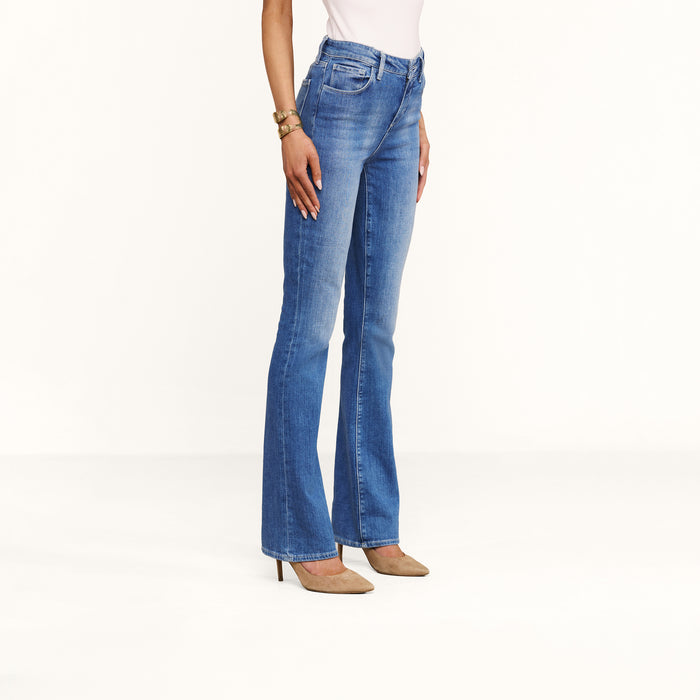 Wearing the Selma Bootcut Jean in a medium wash stretch denim, paired with a white top, tan heels, and a gold bracelet, the woman is shown from the waist down against a plain white background.