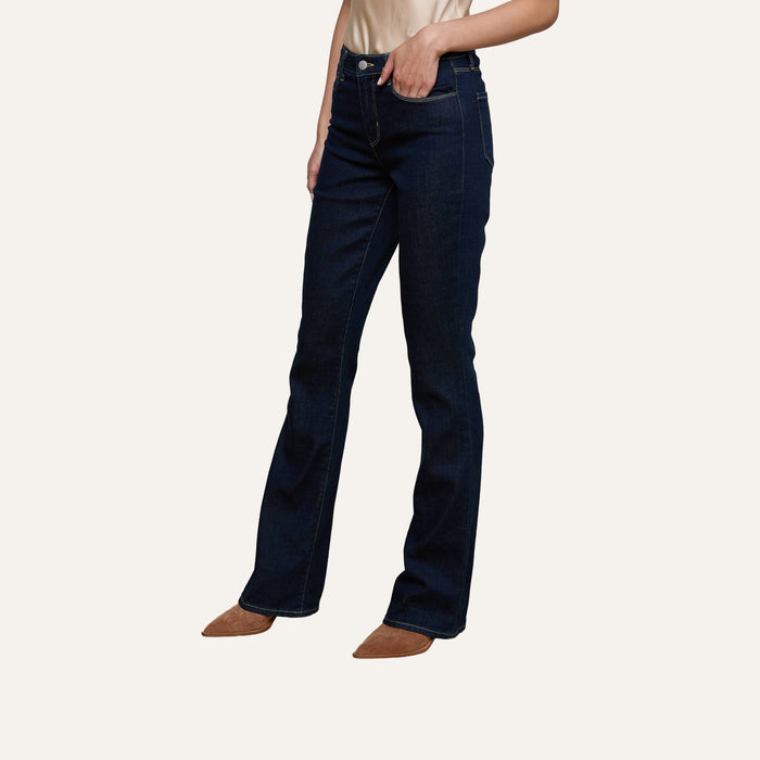 A person wearing the Selma Bootcut Jean in dark blue stretch denim and brown pointed-toe shoes stands against a plain white background, with only the lower half of the body visible.