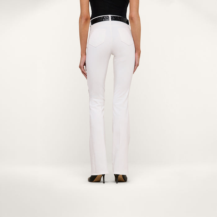 Wearing the Selma Bootcut Jean crafted from stretch denim, a person stands facing away against a plain white background, showcasing the contoured waistband and fit at the back. They style the white jeans with a black top and black high heels.