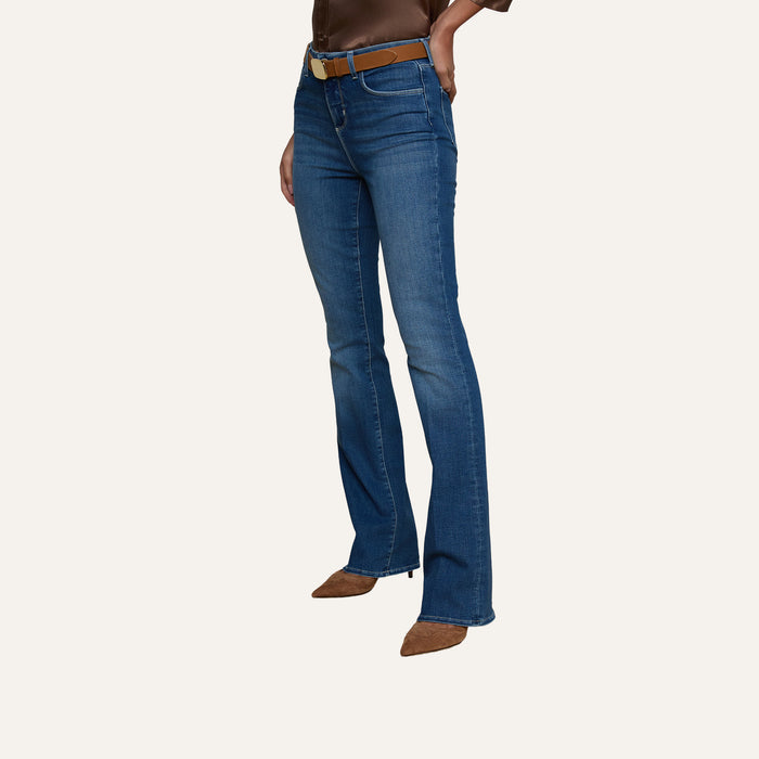 A person is wearing the Selma Bootcut Jean in blue, crafted from super stretch denim, paired with a brown belt and brown pointed-toe shoes. The upper body is partially visible with arms bent at the elbows, set against a plain light-colored background.