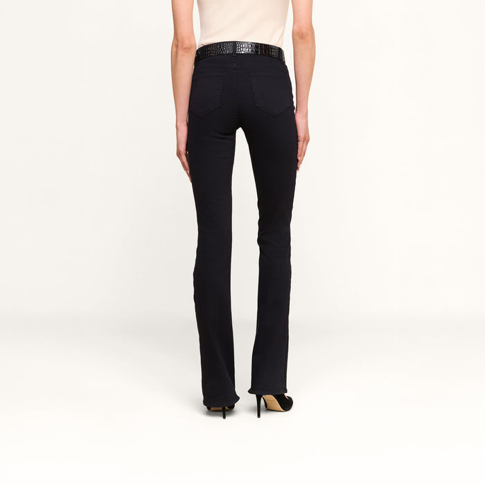 Wearing the Selma Bootcut Jean in a true-black, high-rise silhouette crafted from super stretch fabric, along with a cream sleeveless top and high-heeled shoes, a person stands facing away from the camera against a plain white background.