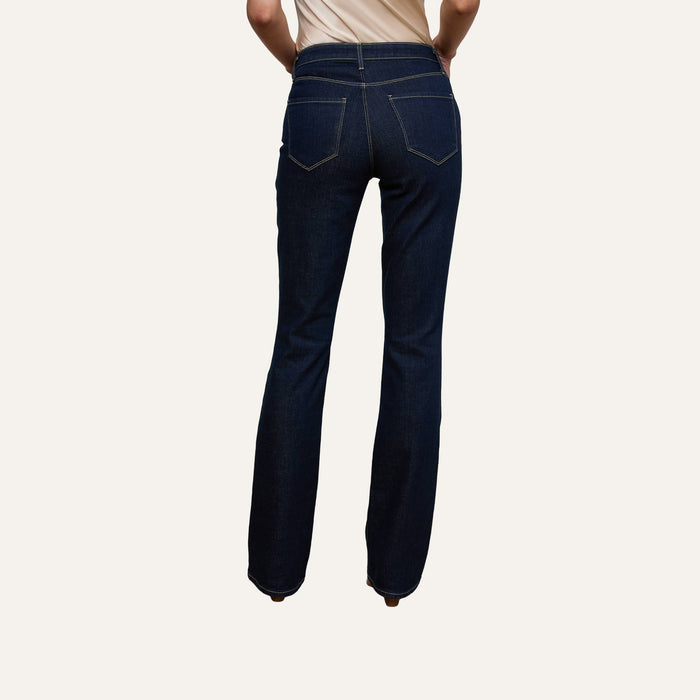 Wearing the Selma Bootcut Jean in dark blue stretch denim paired with a beige top, the person stands with their back to the camera against a plain white background, showcasing the high-rise fit and detailed back pockets of the jeans.