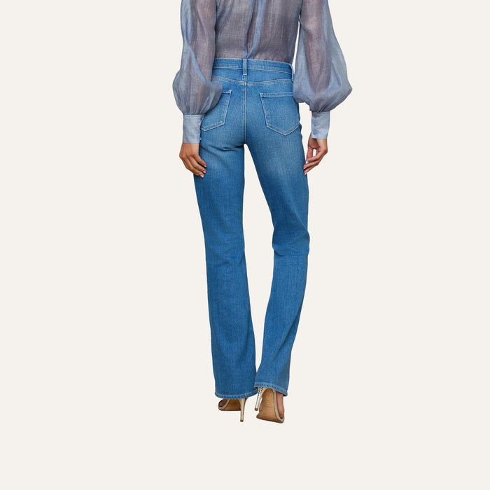 A person stands facing away against a plain, light background, wearing a sheer long-sleeve blouse, the Selma Bootcut Jean in blue featuring a contoured high-rise waistband, and beige heels, showcasing the back of the outfit.