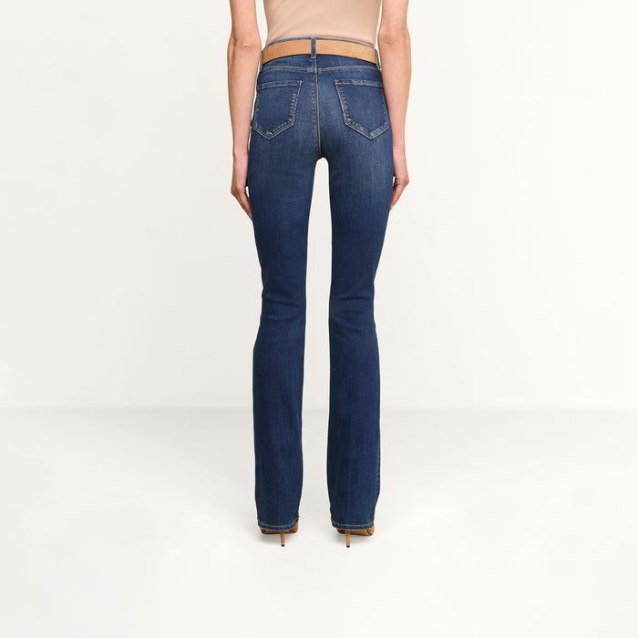 A person is standing facing away, wearing a beige sleeveless top, beige heels, and the Selma Bootcut Jean—a pair of dark blue high-rise slim bootcut jeans crafted from stretch denim—against a plain white background.