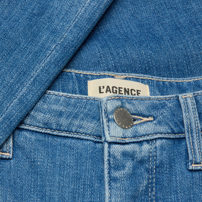 Close-up of the Selma Bootcut Jean in blue denim, showcasing a metal button, contoured waistband, belt loop, and an interior clothing label with the LAGENCE brand name. A denim shirt is partially draped over the jeans.