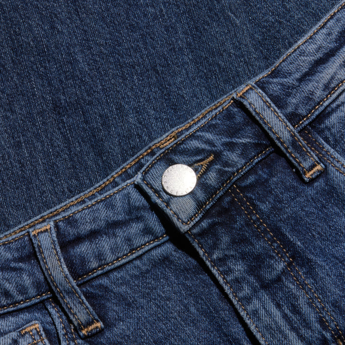 A close-up of blue denim jeans highlights the waistband, metal button, belt loops, and visible stitching against a coordinating denim background.