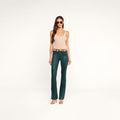 A woman wearing sunglasses, a beige tank top, a brown belt, the Selma Coated Bootcut Jean in teal, and heeled sandals stands against a plain white background.