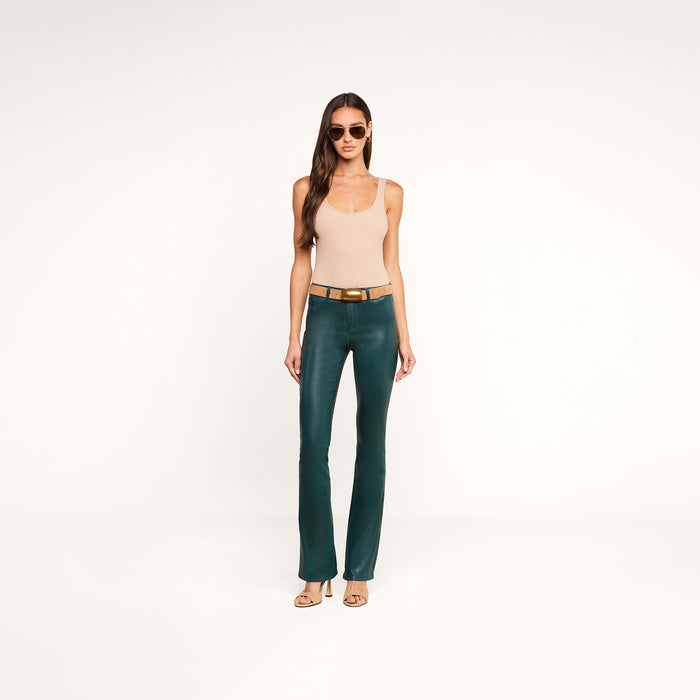 A woman wearing sunglasses, a beige tank top, a brown belt, the Selma Coated Bootcut Jean in teal, and heeled sandals stands against a plain white background.