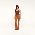 A woman wearing sunglasses, a dark brown Nica Scoopneck Bodysuit, brown leather pants with a belt, and brown shoes stands against a plain white background.