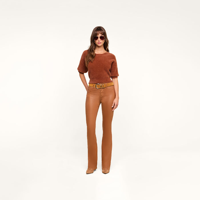 A woman wearing sunglasses, a dark java brown short-sleeve top, tan boots, and the Selma Coated Bootcut Jean stands confidently against a plain white background.