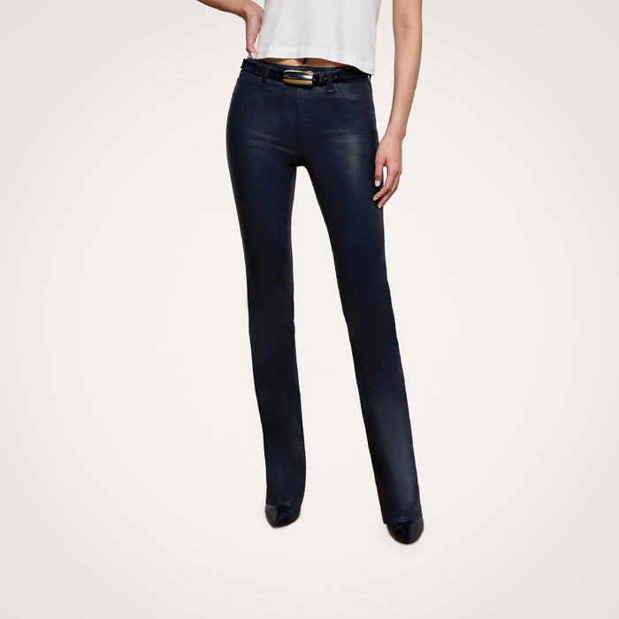 A person wearing a white t-shirt, the Selma Coated Bootcut Jean in a high-rise baby bootcut style, and black shoes stands with one hand on their hip against a plain light background.