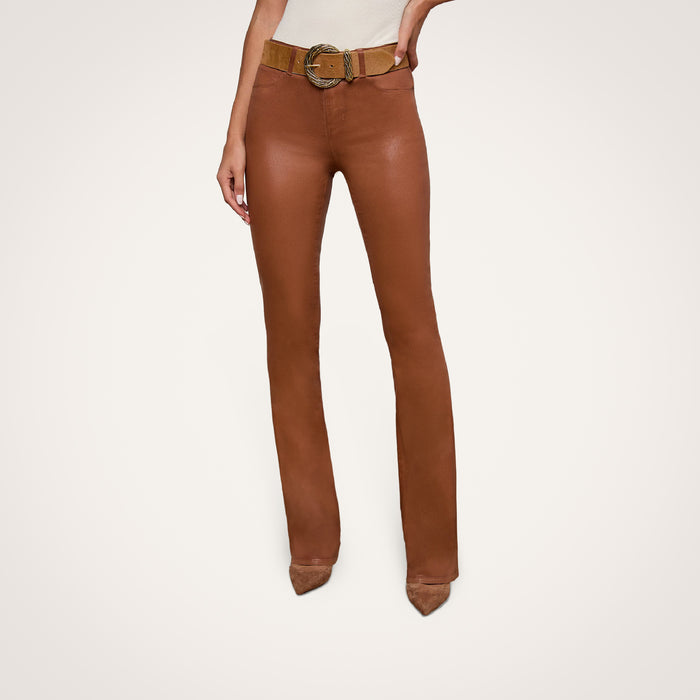 A woman wearing the Selma Coated Bootcut Jean, styled with a wide belt and beige pointed-toe shoes, stands against a plain white background.