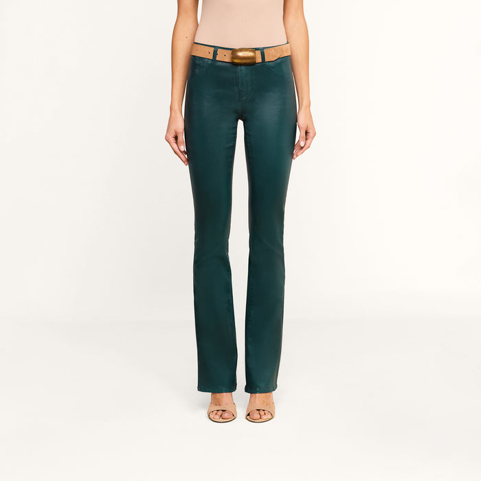 A woman models the Selma Coated Bootcut Jean in dark green, styled with a brown belt and nude heels, standing against a plain white background. Only her body from the shoulders down is shown.