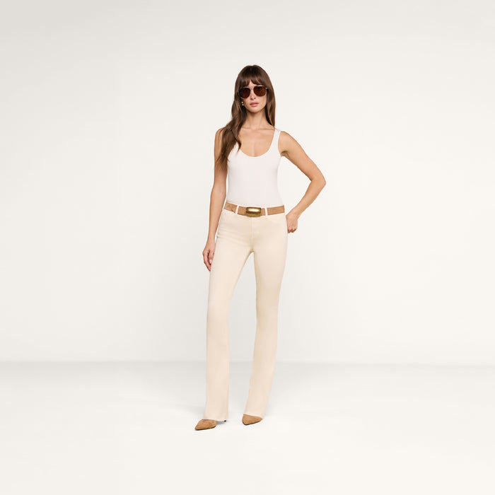A woman wearing sunglasses, a white tank top, and brown shoes stands confidently against a plain white background, pairing her outfit with the Selma Coated Bootcut Jean.