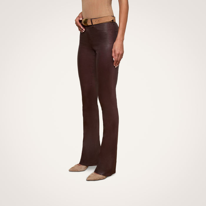 Wearing the Selma Coated Bootcut Jean in chocolate brown, a person with a tan belt and tan pointed-toe shoes stands against a plain white background.