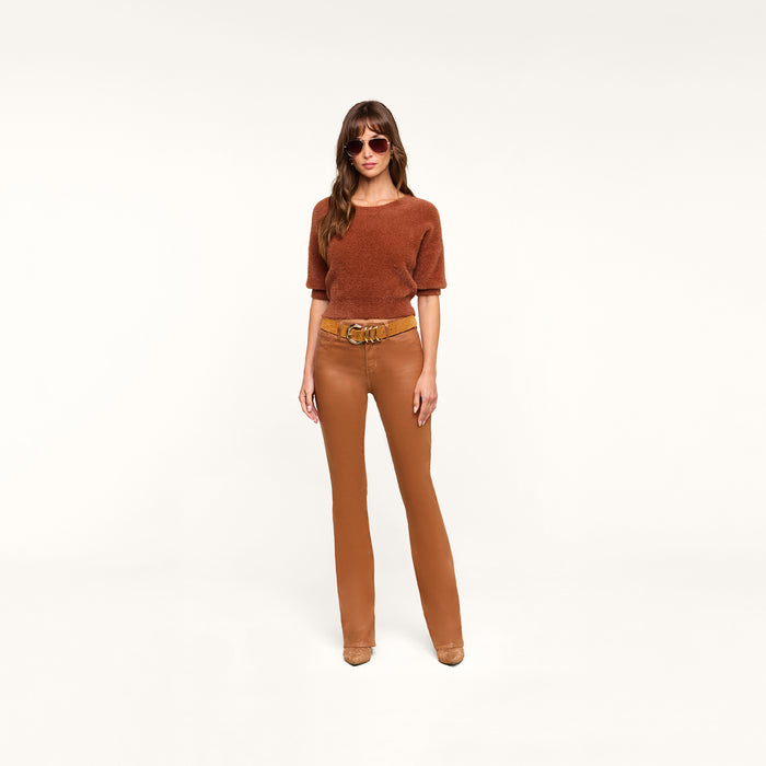 A woman wearing sunglasses, a dark java brown short-sleeve sweater, the Selma Coated Bootcut Jean with a belt, and brown shoes stands against a plain white background.