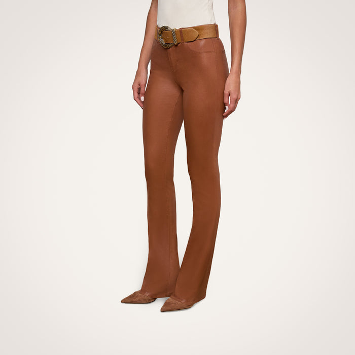 A person wearing the Selma Coated Bootcut Jean, styled with a tan belt and pointed-toe shoes, stands against a plain white background. Only the lower half of their body is visible.