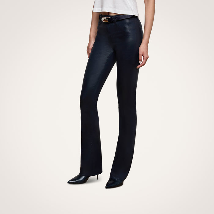 A person wearing the Selma Coated Bootcut Jean, paired with a white top and black pointed-toe shoes, stands against a plain white background. Only the lower half of the body is visible.