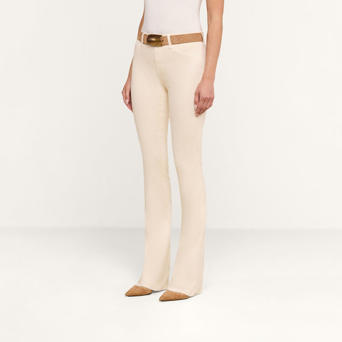 A person wearing the Selma Coated Bootcut Jean in beige, styled with a brown belt, a white top, and brown pointed shoes. They are standing against a plain white background, with their face not visible in the image.