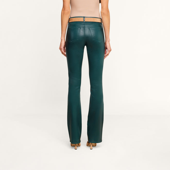 A woman stands facing away, wearing a beige sleeveless top and the Selma Coated Bootcut Jean in dark green with tan heels, against a plain white background.