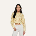 A woman wearing the Seychelle Cropped Eyelet Blouse—a light yellow, long-sleeve poplin lace crop top with a scalloped hem—paired with high-waisted white pants, stands against a plain background.