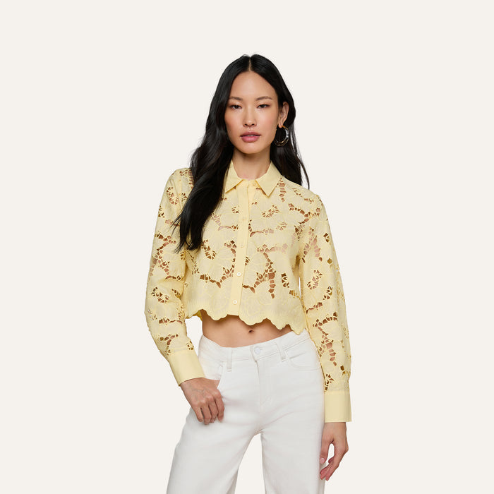 A woman wearing the Seychelle Cropped Eyelet Blouse—a light yellow, long-sleeve poplin lace crop top with a scalloped hem—paired with high-waisted white pants, stands against a plain background.