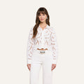 A woman is wearing the Seychelle Cropped Eyelet Blouse—a long-sleeved, white floral lace embroidered top with scalloped edges—paired with high-waisted white belted pants, standing against a plain light background.