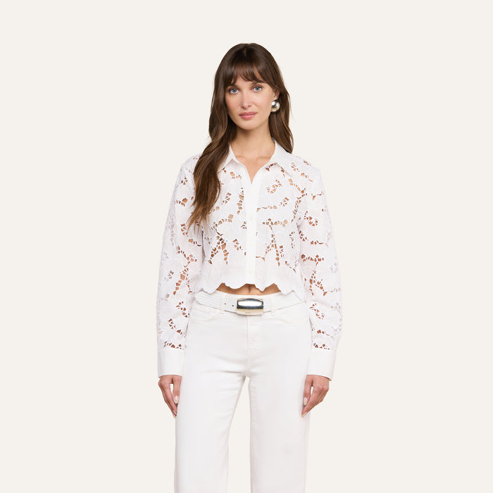 A woman is wearing the Seychelle Cropped Eyelet Blouse—a long-sleeved, white floral lace embroidered top with scalloped edges—paired with high-waisted white belted pants, standing against a plain light background.