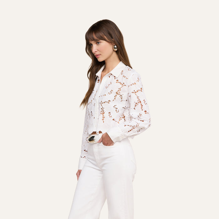 A woman with long brown hair stands in profile against a plain light background, wearing the Seychelle Cropped Eyelet Blouse paired with high-waisted white pants.