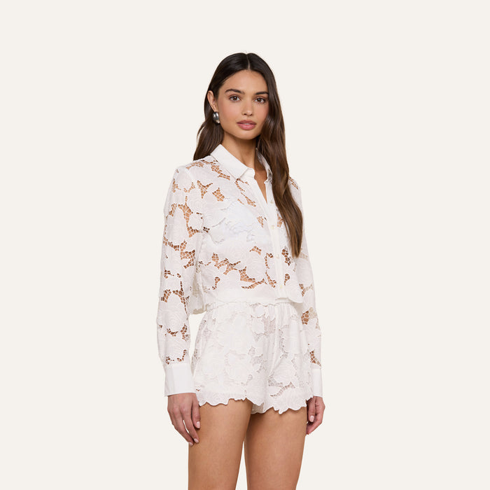 A woman wearing the Seychelle Cropped Eyelet Blouse in white, paired with matching lace shorts, stands against a plain light background, facing slightly to the side.