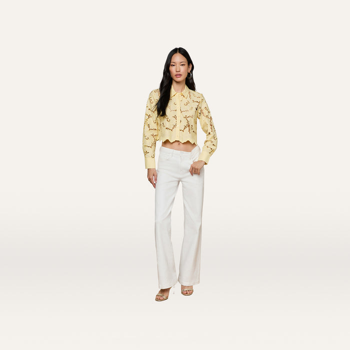 A woman stands against a plain white background wearing the Seychelle Cropped Eyelet Blouse, a yellow long-sleeve eyelet top, paired with white wide-leg pants and heeled sandals.