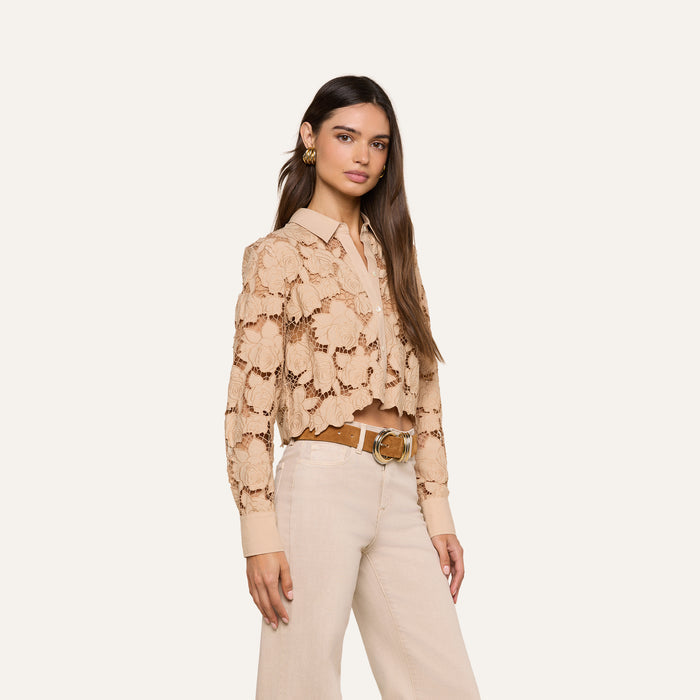 A woman wearing the Seychelle Cropped Eyelet Blouse—a beige floral eyelet top with a cropped silhouette—layered over a collared shirt and paired with high-waisted cream pants and a tan belt, stands against an off-white background.